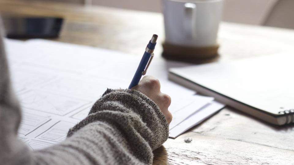 An open worn notebook with a long handwritten checklist beside a laptop showing an online clothing store in warm morning light
